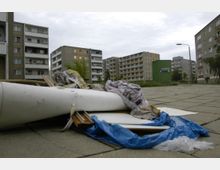 Ansammlung von Sperrmüll, darunter Rohre, Stoffreste und Plastik, auf einer gepflasterten Fläche in einem Wohngebiet mit mehreren Plattenbau-Wohnblöcken im Hintergrund. Rechts steht ein grüner Altkleidercontainer, Bäume und Straßenlaternen ergänzen das urbane Umfeld.