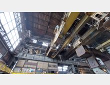 Innenansicht einer großen Industriehalle mit einer hohen Decke aus Stahlträgern und Fenstern, die diffuses Licht hereinlassen. Sichtbar sind gelbe Laufkräne, Metallkonstruktionen sowie verschiedene Plattformen und Kabel, die für industrielle Zwecke genutzt werden.