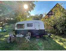 Ein kleiner, weiß-schwarzer Wohnwagen steht auf einer sonnenbeschienenen Wiese unter Bäumen. Davor befinden sich ein runder Tisch mit Geschirr und zwei Stühle, während im Hintergrund ein Fachwerkhaus sichtbar ist.