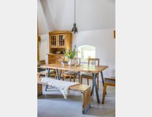 Essbereich in einem Landhausstil mit einem Holztisch, Holzstühlen und einer Holzbank mit Fellauflage. Im Hintergrund steht ein Holzgeschirrschrank, und über dem Tisch hängt eine Metall-Pendelleuchte; ein kleines Fenster sorgt für Tageslicht.