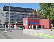 Eingang zur U-Bahn-Station Fehrbelliner Platz in Berlin, mit einem roten Überbau und blauem Stationsschild. Im Hintergrund steht ein modernes Bürogebäude mit großen Glasfassaden und einer Straßenkreuzung mit Ampeln sowie grünen Bäumen.