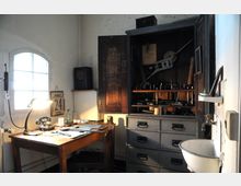 Ein kleines Arbeitszimmer mit einem Holztisch, darauf ein Telefon, eine Schreibtischlampe und verschiedene Papiere. Im Hintergrund ist ein geöffnetes Werkzeugschrank mit großen Werkzeugen zu sehen, daneben eine graue Kommode mit Schubladen und ein kleines Waschbecken an der Wand.