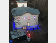 Zweistöckiges Gebäude mit hellem Putz und grauem Metalldach, beleuchtet von farbigen Lichtern (blau und rot) im Außenbereich. Vor dem Eingang befinden sich Stehtische und ein kleiner Vorbau, umgeben von Bäumen in einer abendlichen Umgebung.