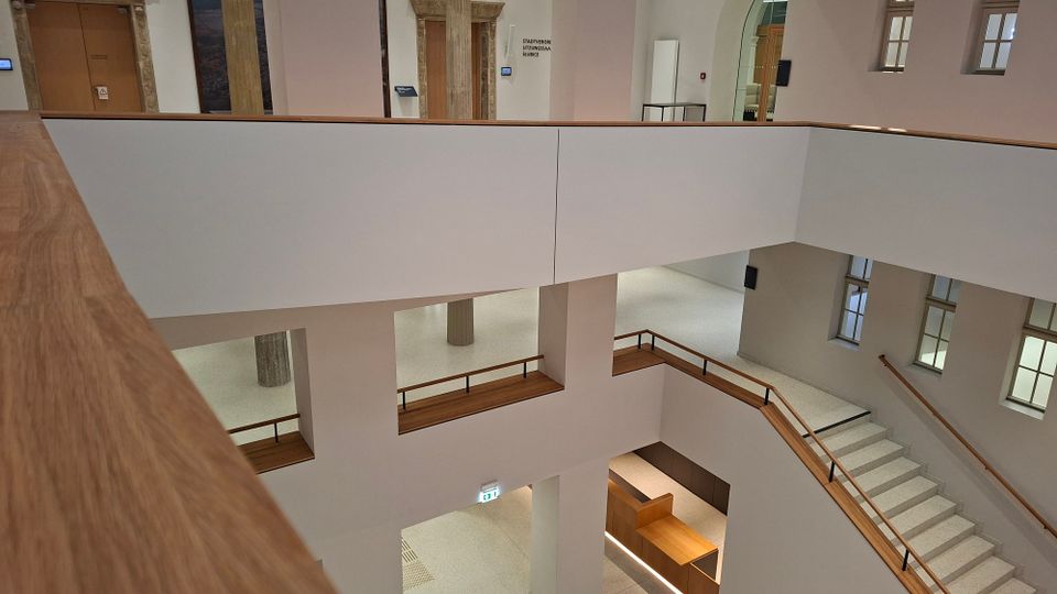 Mehrstöckiges Atrium im Inneren eines öffentlichen Gebäudes mit weißen Wänden, großen Fensteröffnungen und einem Treppenhaus mit Holzhandlauf. Umlaufende Galerien und rechteckige Durchblicke verbinden die Ebenen; unten sind Sitz- und Thekenbereiche aus Holz zu sehen.
