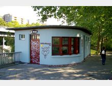 Rundes Gebäude mit hellblauer Fassade und roten Fenstern, dessen Eingangstür mit Graffiti bedeckt ist. Im Hintergrund sind Bäume, ein Weg und teilweise Wohngebäude sichtbar.