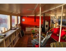 Wohnraum auf einem Hausboot mit Holzboden, roter Wand, großen Fenstern und gemütlicher Einrichtung. Zu den Möbeln gehören ein Regal mit Büchern, ein Kaminofen, ein Tisch mit einem Blumenstrauß und eine rote Couch.