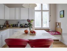 Helle Küche mit weißen Einbauschränken, einer Holzarbeitsplatte und einer Kochinsel mit roten Barhockern. Auf der Insel stehen Obst und eine Vase mit Blumen, im Hintergrund ein großes Fenster mit Blick ins Freie und eine gepolsterte Sitzbank an der Wand.