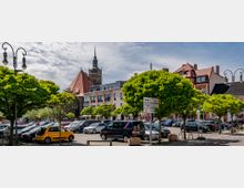 Stadtplatz mit geparkten Autos, umgeben von Bäumen und historischen Gebäuden, darunter eine Kirche mit einem hohen Turm und einer Uhr. Im Hintergrund moderne und traditionelle Fassaden, Straßenschilder und Laternen im Vordergrund.