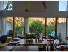 Wohnzimmeransicht mit großer Fensterfront, die auf einen begrünten Garten und eine blaue Gartenbank zeigt. Im Innenraum stehen ein Holztisch mit Stühlen, darauf Blumen, Geschirr und Gewürzflaschen; im Hintergrund eine Couch mit Kissen und ein Wandbild.