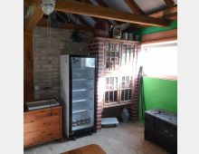 Rustikaler Innenraum mit einer teils unverputzten Ziegelwand. Zu sehen sind ein freistehender Kühlschrank, eine Holzvitrine mit Glastüren, ein kleiner Holzofen und Holzbalkendecken. An der rechten Seite befindet sich eine grüne Wand mit einem Fenster.