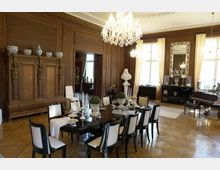 Elegantes Esszimmer mit Holzwänden, einem großen Esstisch mit weißen Stühlen und dekorativem Gedeck. Ein Kronleuchter hängt von der Decke, und große Fenster mit hellen Vorhängen lassen viel Tageslicht herein; ein Klavier und Spiegel ergänzen den luxuriösen Raum.