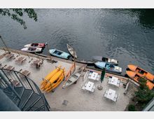 Blick von oben auf eine Uferpromenade mit Sandboden, auf der mehrere weiße Tische und Stühle stehen. Am Wasser sind verschiedene Boote und Kanus vertäut, darunter gelbe Kajaks und ein orangefarbenes Motorboot. Eine Wendeltreppe ist links im Bild sichtbar.
