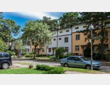 Reihenhäuser mit pastellfarbigen Fassaden in einer ruhigen Wohnsiedlung, umgeben von Bäumen und Grünflächen. Im Vordergrund sind eine gepflasterte Straße, parkende Autos, Fahrräder und eine Straßenlaterne mit einem Straßenschild zu sehen.