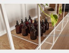 Regal mit weißen Metallrahmen, gefüllt mit braunen Sprayflaschen auf einer hölzernen Ablage; daneben ein geflochtener Korb und eine Pflanze mit grünen Blättern.