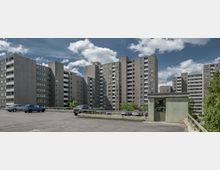 Mehrere graue Hochhäuser in Plattenbauweise, die sich um einen offenen Bereich mit aufgestellten Autos erstrecken. Im Vordergrund eine mit Graffiti versehene Mauer und ein Parkplatz; Bäume grenzen den Bereich ab.