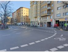 Straßenszene mit Kreuzung in einer Stadt, umgeben von mehrstöckigen Wohn- und Geschäftshäusern mit gelblicher Fassade. Im Vordergrund ein Fahrradweg und Markierungen auf der Straße, rechts ein Gebäudeeingang mit der Beschilderung „Kaiser-Apotheke“ und parkende Fahrräder.