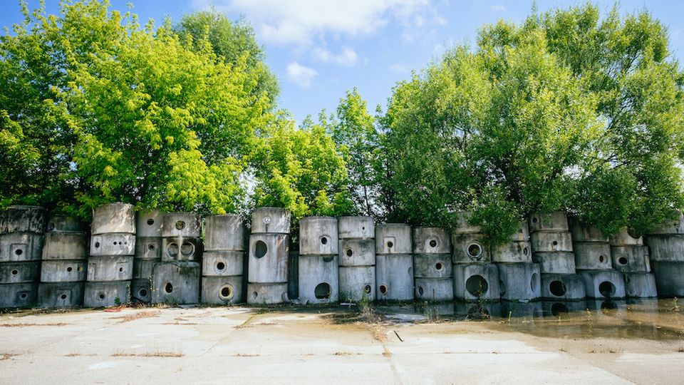Stapel aus Betonblöcken mit runden Öffnungen vor einer Reihe dicht belaubter Bäume, auf einem betonierten, teils mit Wasser überschwemmten Platz unter einem blauen Himmel.