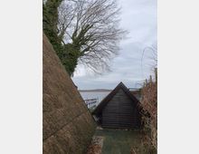 Reetgedeckte Gebäude und ein Holzschuppen stehen nahe einem Ufer. Im Hintergrund ist ein See mit einem schmalen Steg, kahlen Bäumen und einer bewölkten Himmel zu sehen.
