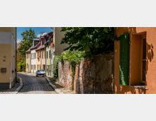 Schmale, gepflasterte Gasse mit farbenfrohen, dicht aneinandergereihten Wohnhäusern mit Blumen an den Fenstern; rechts eine alte, bewachsene Ziegelmauer und ein Haus mit orangefarbener Fassade und grünem Fensterladen. Im Hintergrund stehen Bäume und ein parkendes Auto ist sichtbar.