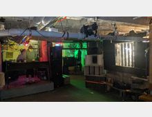 Ein dunkler Kellerraum mit niedriger Decke, sichtbaren Rohren und Kabeln an der Decke. Links ist ein DJ-Pult mit einem DJ und bunter Beleuchtung, im Hintergrund sind Lautsprecher, Regale und technische Geräte zu erkennen.