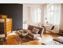 Helles Wohnzimmer mit Parkettboden, einem großen goldenen Kachelofen links, einem grauen Sofa, einem dekorativen Teppich und einem modernen schwarzen Stuhl. Rechts steht ein kleiner Esstisch mit zwei Stühlen vor einem großen Fenster mit weißen Vorhängen.