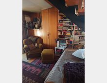 Gemütlicher Wohnraum mit Holzverkleidung, einem großen Bücherregal voller Bücher, einem gelben Sessel mit passendem Fußhocker und einem Bett im Vordergrund. Eine schwarze Treppe führt nach oben, und ein bunter Teppich bedeckt den Boden.