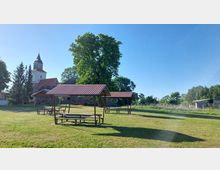 Grüne Wiese mit drei überdachten Picknicktischen im Vordergrund. Im Hintergrund steht eine Kirche mit Kirchturm sowie weitere Gebäude, umgeben von Bäumen und freier Landschaft bei klarem Himmel.