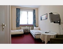 Kleines Hotelzimmer mit zwei Einzelbetten, einem runden Tisch mit weißer Tischdecke, einem Glas und Fernbedienungen. Ein Fenster mit blau gemusterten Vorhängen erhellt den Raum, der mit rotem Teppichboden ausgelegt ist. Ein Röhrenfernseher ist an der Wand montiert.