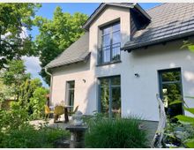 Modernes Einfamilienhaus mit hellgrauer Fassade, großen Fenstern und einem Dach aus Schieferziegeln. Im Vordergrund ein Garten mit Pflanzen und eine Terrasse mit Tisch und Stühlen, umgeben von Bäumen und Grünflächen.