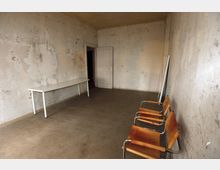 Ein schlichter Raum mit einer verputzten, teils abblätternden Wand, einem Betonboden und einer schmalen weißen Tür im Hintergrund. Es stehen ein schmaler weißer Tisch an der linken Wand und mehrere orange-braune Stühle mit Metallrahmen an der rechten Wand.