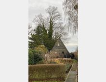 Ein kleines, dunkles A-förmiges Holzhaus mit grün gerahmten Fenstern, umgeben von einem Garten mit kahlen Sträuchern und Hecken; im Hintergrund stehen hohe, teils kahle Bäume unter bewölktem Himmel.