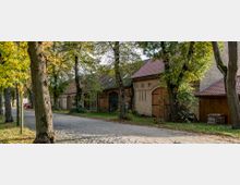 Eine schmale, von Bäumen gesäumte Kopfsteinpflasterstraße führt an einer Reihe historischer Backsteingebäude mit roten Ziegeldächern und Holztoren entlang. Im Vordergrund stehen herbstlich belaubte Bäume, die den Weg teilweise beschatten.