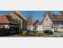 Kleiner Dorfplatz mit historischen Fachwerkhäusern, gepflasterten Straßen und einem zentralen Blumenbeet; im Vordergrund ein geparkter Pkw und ein Baum, der noch keine Blätter trägt.