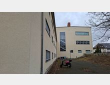 Hof zwischen zwei hell verputzten Mehrgeschossgebäuden mit vielen rechteckigen Fenstern und einem hohen, verglasten Treppenhaus. Der asphaltierte Platz ist von Laub bedeckt; am Rand stehen eine Laterne und eine niedrige Steinmauer mit angrenzenden Wohnhäusern im Hintergrund.