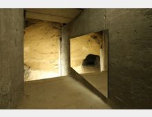 Betonraum mit einer Wandöffnung, die einen Blick in eine natürliche, sandsteinartige Höhlenformation ermöglicht; ein großer Spiegel an der rechten Wand reflektiert einen Teil der Höhle.