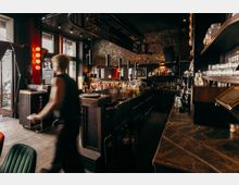 Innenraum einer Bar mit langer dunkler Theke, Regalen voller Gläser und Flaschen sowie warmem Licht. Backsteinwand, schwarze Decke mit Industrieelementen und große Fenster zur Straße prägen den Raum.
