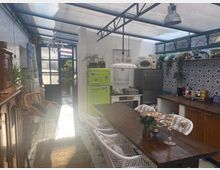 Ein heller Innenraum mit Küche, der durch ein transparentes Dach beleuchtet wird. Es gibt einen Holzesstisch mit weißen Stühlen, eine grüne Retro-Kühl-Gefrierkombination, blaue Fliesen mit Mustern an der Rückwand und Pflanzen sowie Küchenutensilien auf Regalen.