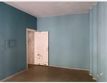 Leerer Raum mit hellblauen Wänden und einer alten weißen Holztür, die teilweise geöffnet ist und in einen angrenzenden, unbehandelten Bereich führt; Betonboden und eine minimalistische Gestaltung sind sichtbar.