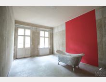 Ein leerer Raum mit Betonboden, abgenutzten Wänden und einem roten Farbakzent an der rechten Wand. Links befinden sich zwei Sprossenfenster mit Glastüren, und im Raum steht eine frei stehende, bunt gemusterte Badewanne mit Löwenfüßen.