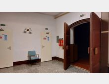 Ein Flur mit grauem Steinboden, zwei weißen Türen und einer breiten, offenen Tür mit braunen, gepolsterten Flügeln. An der Wand hängen zwei Feuerlöscher, und unter einer Wandlampe steht ein einfacher Stuhl mit türkisfarbenem Polster.