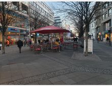 Eine städtische Fußgängerzone mit Geschäften und Restaurants auf beiden Seiten, teils mit großen Glasfassaden. Im Vordergrund stehen Tische und Stühle eines Straßencafés unter roten Sonnenschirmen, während Passanten und Radfahrer die Straße beleben.