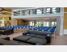 Ein moderner Gemeindesaal mit Stuhlreihen in blauem Polster, einer zentralen Holzkanzel und großen Fenstern im Hintergrund, die Licht hereinlassen. Im Vordergrund befinden sich schwarze Ledersofas und mehrere aufgeschlagene Bücher.