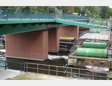 Grüne Stahlbrücke mit gemauerten Pfeilern über einen Wasserkanal, neben dem sich technische Anlagen mit grünen Tanks und Geländern befinden. Im Vordergrund liegt ein kleines Rettungsboot am Ufer.