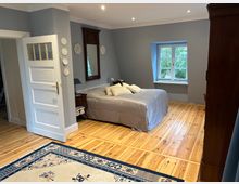Schlafzimmer mit hellem Holzdielenboden, grauen Wänden und einem Doppelbett mit grauer Tagesdecke; daneben stehen ein Nachttisch und ein hoher Spiegel. Ein weißes Sprossenfenster mit Blick ins Grüne sowie eine offene weiße Tür und dunkle Holzschränke rahmen den Raum.