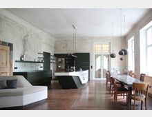 Offene Wohnküche mit industriellem Design: Eine moderne Küche mit dunklen Schränken und einer weißen freistehenden Kücheninsel steht im Vordergrund, während rechts ein langer Holztisch mit unterschiedlichen Stühlen und hängenden Lampen zu sehen ist; die Wände wirken roh und unverputzt. Große Fenster lassen viel Tageslicht in den Raum.