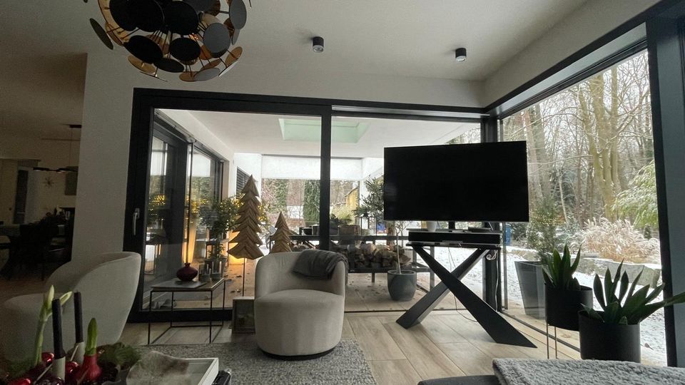 Moderner Wohnbereich mit großer Glasschiebetür und bodentiefen Fenstern, die auf eine überdachte Terrasse und einen verschneiten Garten mit Bäumen hinausgehen. Im Raum stehen ein Sessel auf Teppich, ein Fernseher auf einem X-förmigen Tisch sowie Pflanzen und eine markante Deckenleuchte.