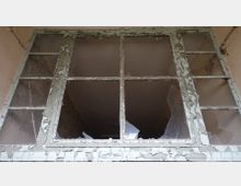 Ein großes, vierteiliges Fenster in einem verfallenen Gebäude, mit abgeblätterter Farbe am Rahmen und mehreren zerbrochenen Glasscheiben. Dahinter sind ein leerer, dunkler Raum und verputzte, teils beschädigte Wände zu sehen.