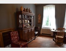 Ein historisch eingerichteter Raum mit Holzboden, einer verzierten Anrichte aus Holz, auf der Keramik und Porzellan stehen, sowie einem großen Fenster mit weißen Vorhängen. Im Vordergrund stehen ein gepolsterter Stuhl und ein Holztisch mit Stuhl.