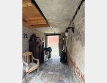 Schmaler Gang mit einem Boden aus unregelmäßig verlegten Ziegelsteinen, einer rohen Beton-Decke und Wänden aus teils verputzten Ziegeln. Links stehen ein rustikaler Holzstuhl und eine dunkle Holzkommode, rechts befinden sich verschiedene Gegenstände, darunter Lampen und Säcke, mit Blick auf einen gepflasterten Hof durch den offenen Durchgang.