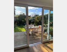 Blick durch eine gläserne Schiebetür auf eine Holzterrasse mit einem Tisch und Sitzbänken, die mit Geschirr und Dekoration gedeckt sind, dahinter ein gepflegter Garten mit Bäumen, Sträuchern und Rasen unter einem sonnigen Himmel.